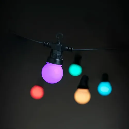 Viedā gaismas ķēde ar apaļām spuldzītēm uz melna kabeļa; fokusā tuvplāns uz violetu spīdumu, fonā izplūdušas daudzkrāsainas gaismas.