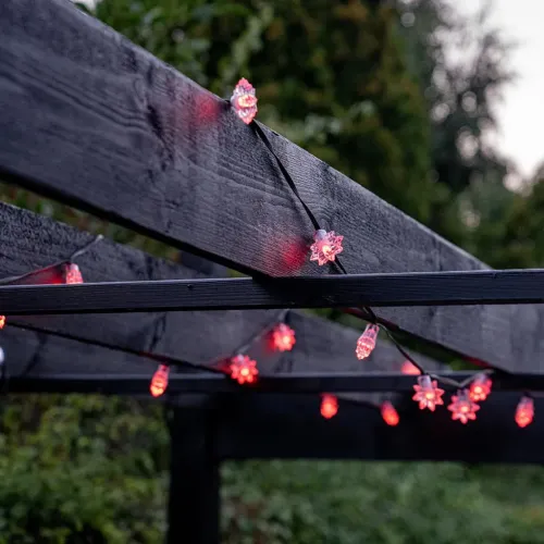 Gaismas ķēde ar zvaigznes formas LED spīd sarkanā krāsā un ir piekarināta uz melnas koka lapenes ārā, krēslas laikā, gar sijām.