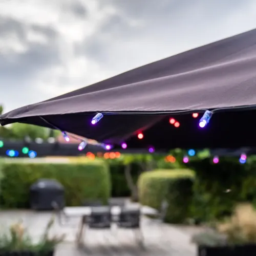 Nutikas valguskett väikeste mitmevärviliste valgusdioodidega piki tumeda päikesevarju serva õues aias, mille taustal on terrass.