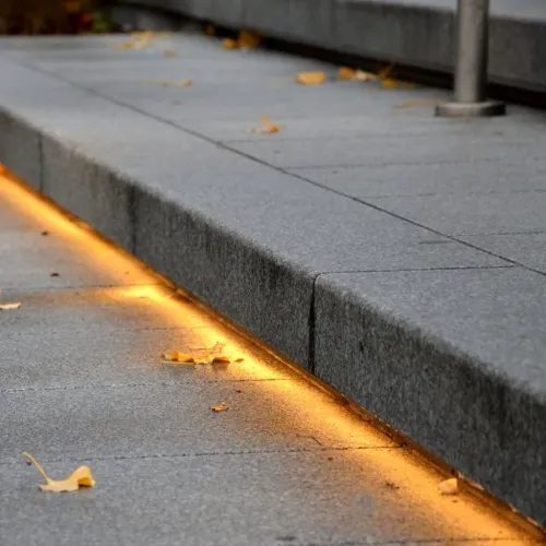 LED sloksne padziļinātā gropē gar āra kāpnēm spīd silti dzeltenā krāsā, iezīmējot malas līniju starp pelēkām lielām akmens plāksnēm.