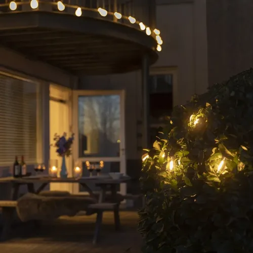 Gaismas ķēde ar senlaicīgām Edisona spuldzēm silti balti spīd, piekārta gar balkona margām un saulrietā ievīta krūmā uz terases.