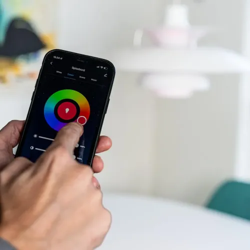 Lite Bulb Moments smarta glödlampor styrs i app: hand justerar färghjul på mobil, med vit pendellampa suddig i bakgrunden.