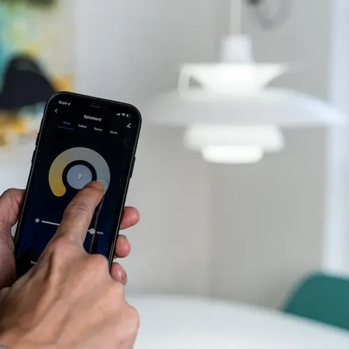 Išmaniosios lemputės "Lite Bulb Moments" valdomos išmaniuoju telefonu; viena ranka programėlėje reguliuojama šviesa, o už jos šviečia balta švytuoklė.